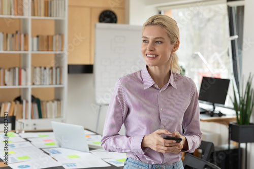 Close up dreamy smiling businesswoman holding smartphone  looking to aside  standing in modern office room  happy young woman entrepreneur employee visualizing future  pondering project strategy