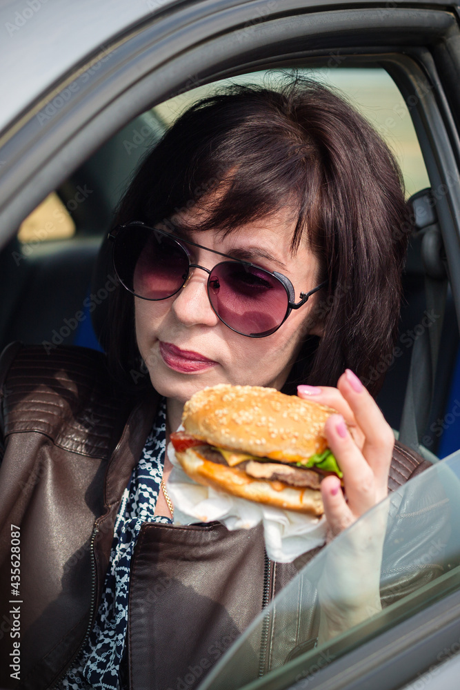 A woman in sunglasses at the wheel of car eats a hamburger. A woman in sunglasses at the wheel of car eats a hamburger.