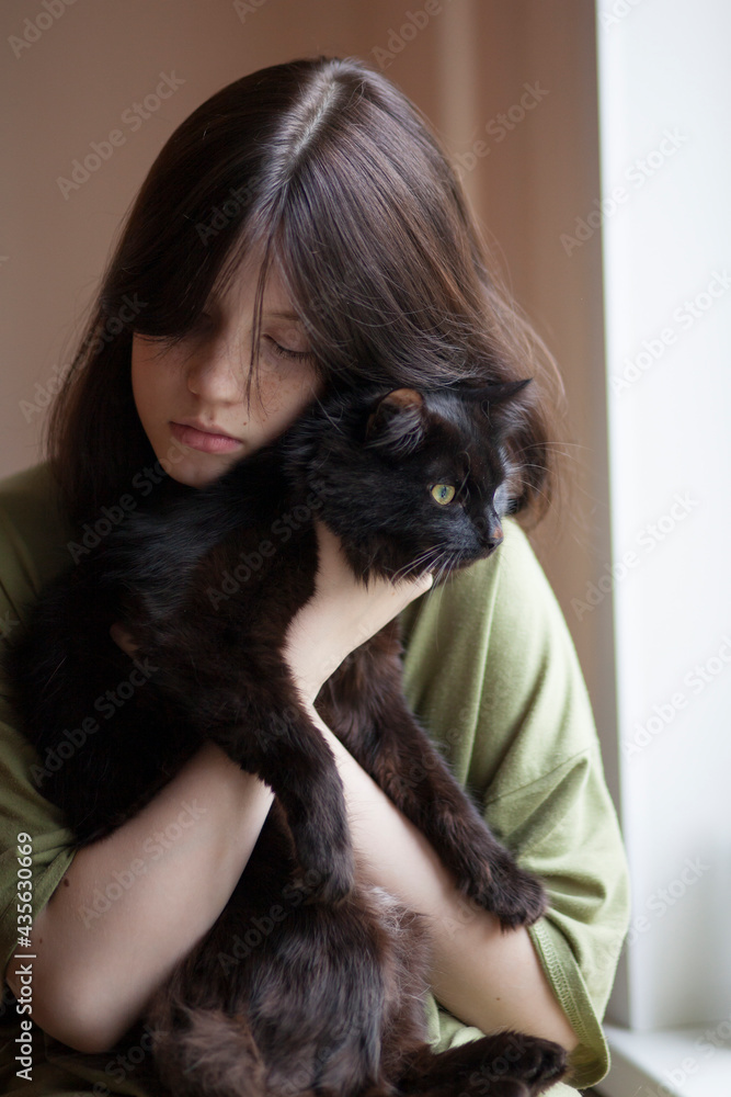 A teenage girl holds in her arms, hugs and kisses a black cat in an apartment.