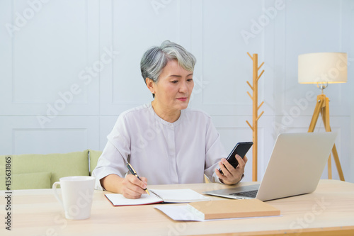 Senior Asia woman working from home using laptop and checking message on smart phone.