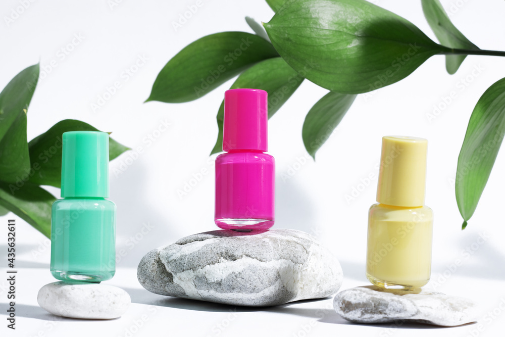 A collection of colored nail manicure polishes on the podium, on a white background, with hard shadows and leaves. Creative mockup of isolated cosmetic bottle with yellow, green and pink nail polish.