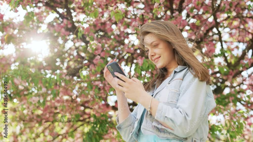 Hipster woman typing by mobile phone outdoors. Cheerful girl with smartphone in park on a background of sakura trees. Smiling lady holding cellphone in hands outside. Hipster woman typing by mobile phone outdoors. Cheerful girl with smartphone in park on a background of sakura trees. Smiling lady holding cellphone in hands outside.