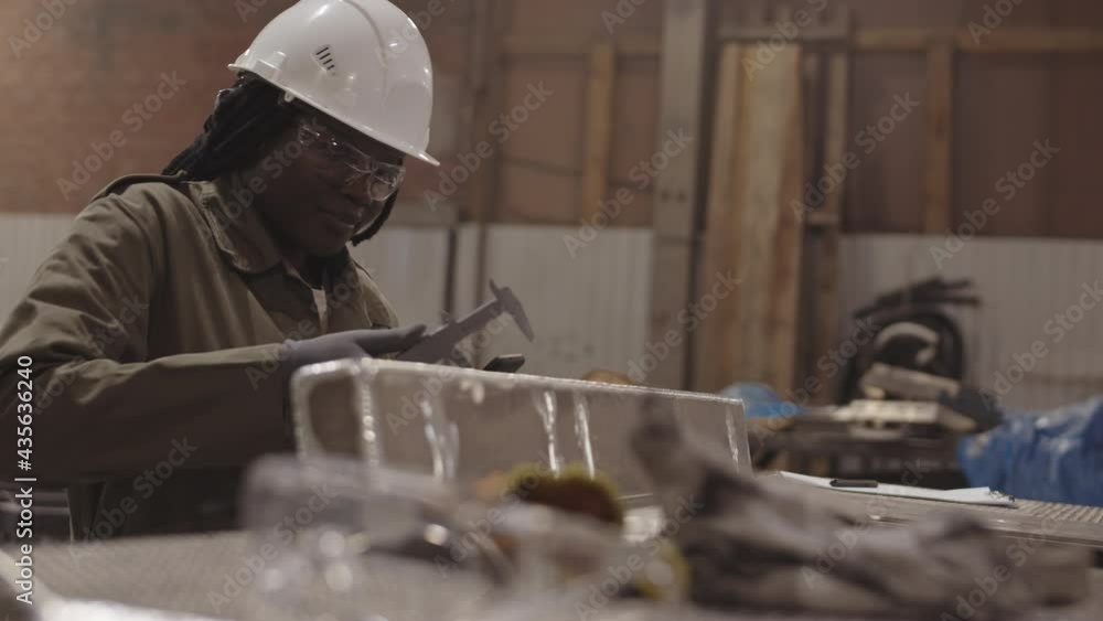 Medium close-up of female African worker wearing white hard hat and safety goggles, using caliper measuring width of metallic piece, then reconciling dimensions on phone Medium close-up of female African worker wearing white hard hat and safety goggles, using caliper measuring width of metallic piece, then reconciling dimensions on phone