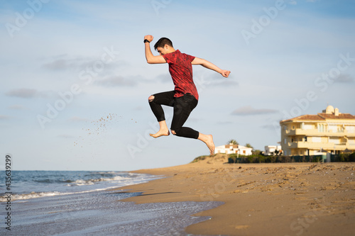 Man jumping on the seashore on a sunny day. Caucasian with long trousers and short-sleeved shirt. Shadow can be seen on the golden sand.
