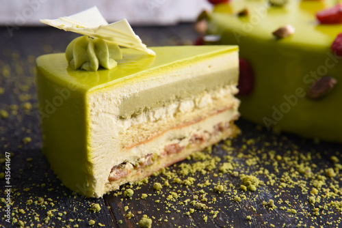 Green glaze cake decorated with pistachios and edible foil on a black wooden background. A slice of cheesecake next to a large pie..