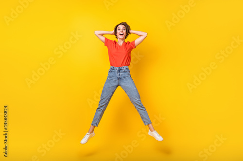 Full size photo of cool brunette optimistic lady jump hands head wear red t-shirt jeans sneakers isolated on yellow color background