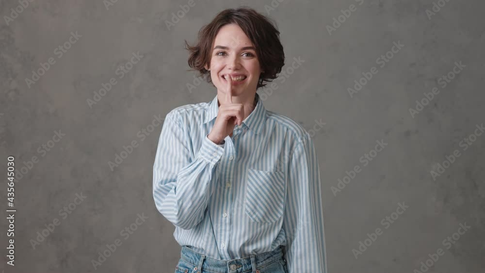 The short-haired happy woman showing a silence gesture and looking to the sides while standing in a gray studio The short-haired happy woman showing a silence gesture and looking to the sides while standing in a gray studio