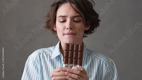 A close-up view of a woman with short hair holding a chocolate bar in her hands and biting off a piece with pleasure in a gray studio