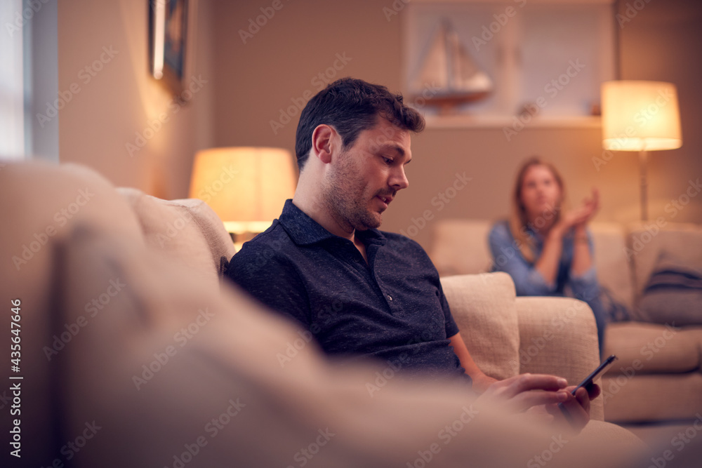 Evening Shot Of Couple On Sofa At Home With Man Looking At Mobile Phone And Unhappy Woman Behind Evening Shot Of Couple On Sofa At Home With Man Looking At Mobile Phone And Unhappy Woman Behind