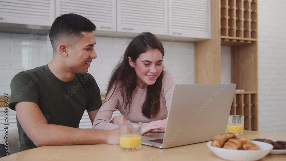 The smiling couple looking for something on a laptop and waving at the camera at the breakfast while sitting in the kitchen The smiling couple looking for something on a laptop and waving at the camera at the breakfast while sitting in the kitchen