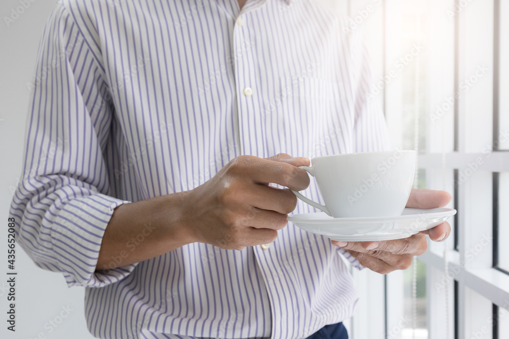 young man standing by the window with a coffee cup at his house in the morning prepares to go to work. Businessman stand to eat coffee in a relaxed mood in the office. young man standing by the window with a coffee cup at his house in the morning prepares to go to work. Businessman stand to eat coffee in a relaxed mood in the office.