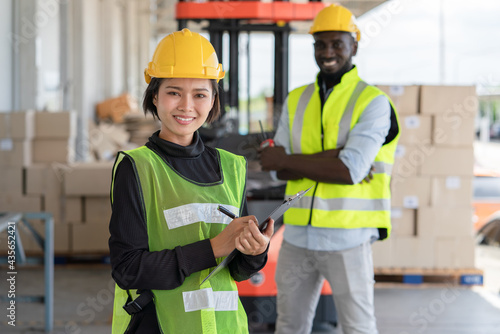 Young Asian woman with man worker in safety vest and yellow helmet working at shipment at warehouse factory