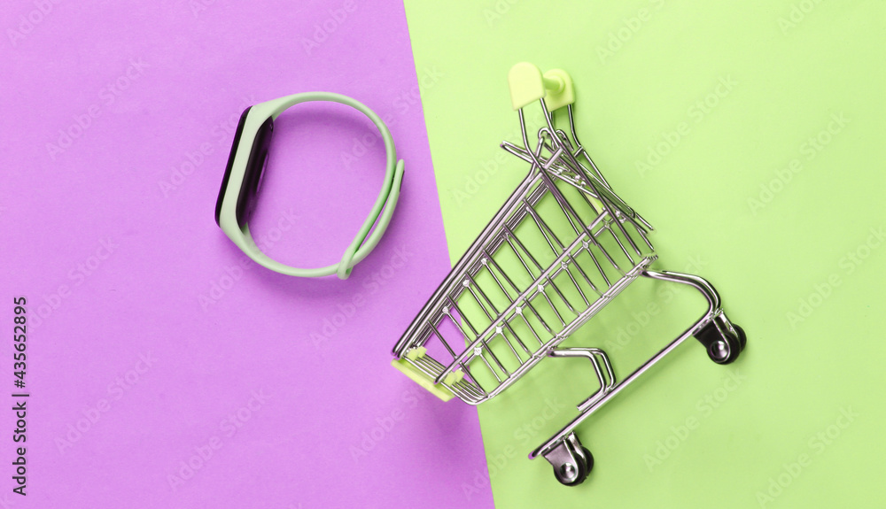Fototapeta premium Mini shopping trolley with smart bracelet on pink green pastel background. Modern gadgets. Top view