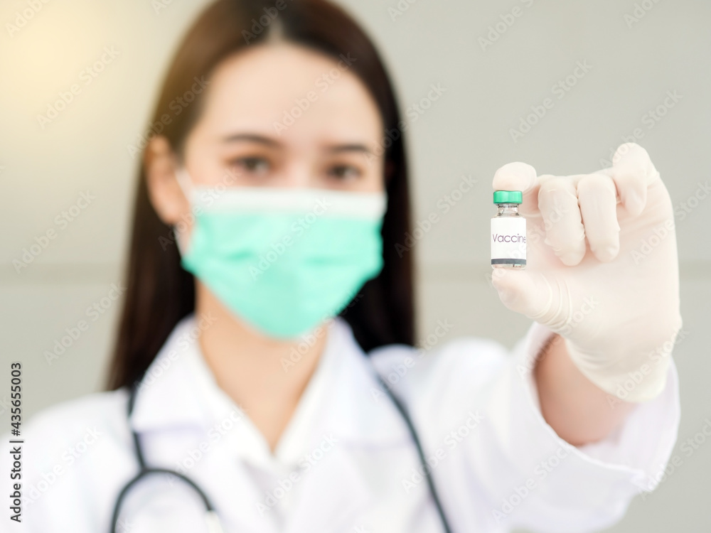 An Asian female doctor or nurse with a surgical mask is holding a vaccine bottle in her hand, focusing on the vaccine bottle in the foreground. An Asian female doctor or nurse with a surgical mask is holding a vaccine bottle in her hand, focusing on the vaccine bottle in the foreground.