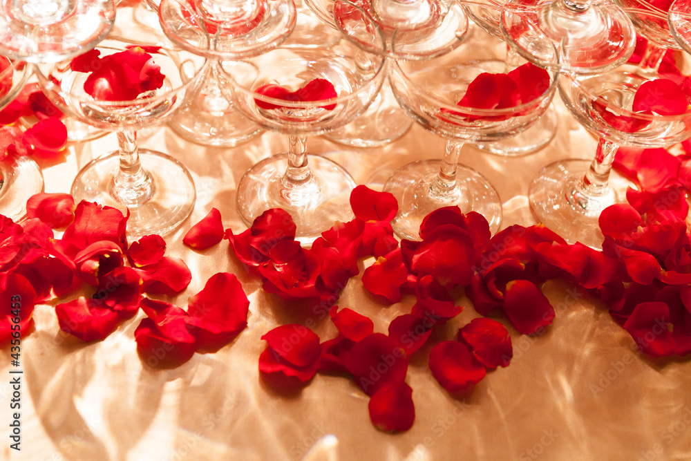 The red rose petals are spilled in and around the wine goblet on the golden banquet table, accompanied by the light reflecting the dazzling light pattern, the perspective taken from top to bottom