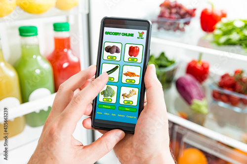 Man using smart phone, buying food. Close-up hand with phone. Grocery shopping online. Mobile app of online food store. Fresh vegetables on fridge shelves. Healthy lifestyle and food delivery.