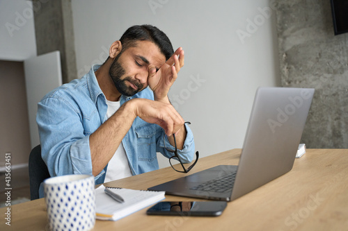 Young indian eastern tired exhausted business man rubbing eyes sitting in modern home office with laptop on desk. Overworked burnout academic Hispanic student with glasses in hand feeling eyestrain. photo