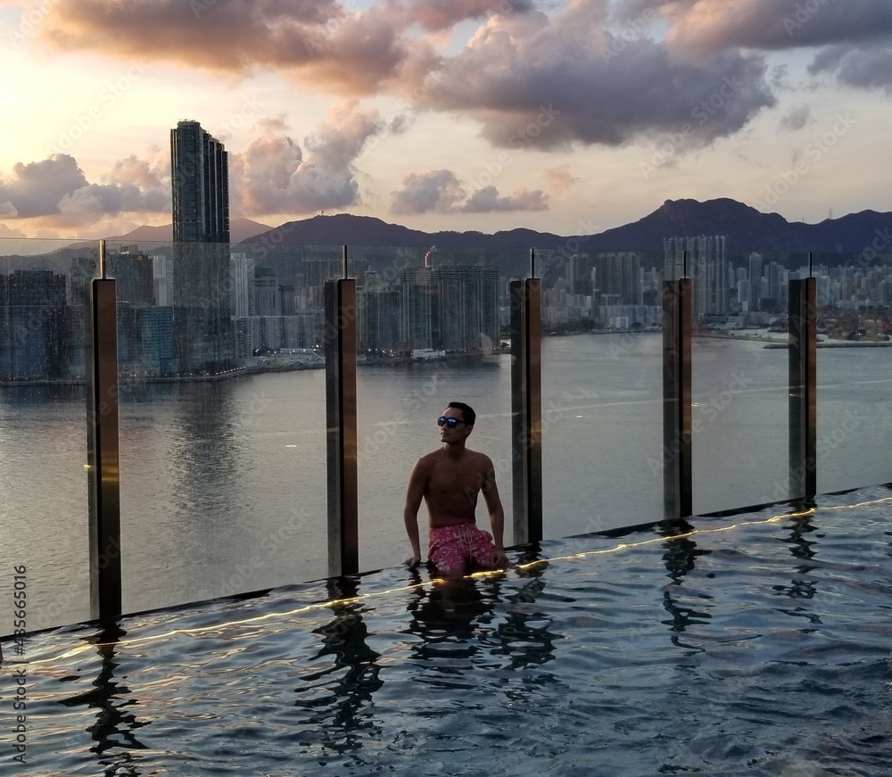 young man relaxing and swimming in a luxury hotel infinity pool overlooking hong kong harbour during a breathtaking beautiful sunset sunrise