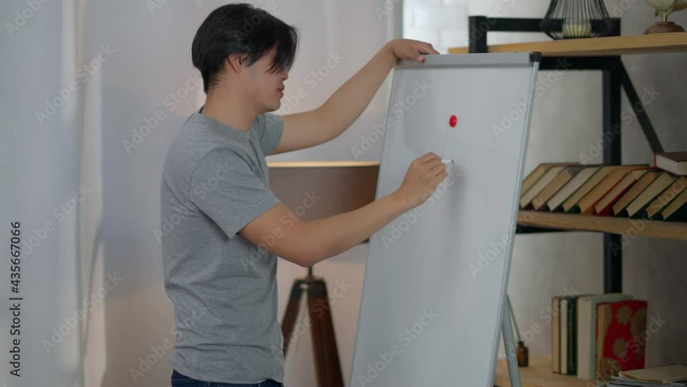 Side view of smiling Asian man drawing on whiteboard standing in home office indoors. Middle shot of cheerful happy millennial planning business work time on remote working Side view of smiling Asian man drawing on whiteboard standing in home office indoors. Middle shot of cheerful happy millennial planning business work time on remote working