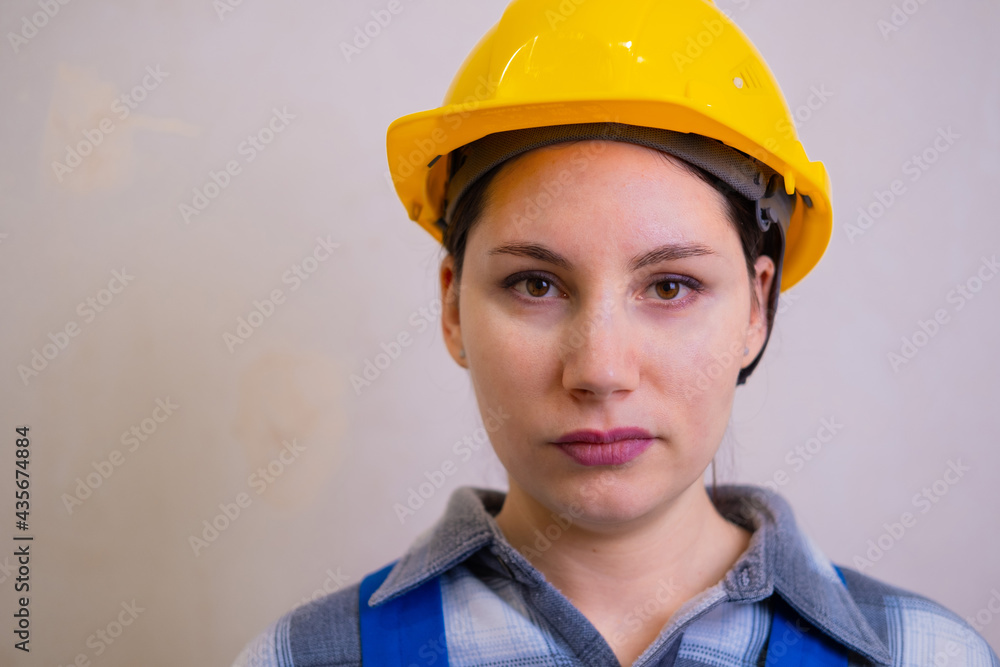 A beautiful young brunette woman in a yellow hard hat and blue uniform looks at the camera on a gray blurred background A beautiful young brunette woman in a yellow hard hat and blue uniform looks at the camera on a gray blurred background