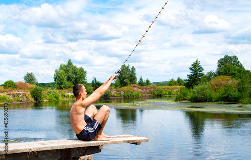 Boy teenager sits on a wooden bridge going to jump into the river from the swinging rope on sunny summer day. Beautiful landscape, copy space.