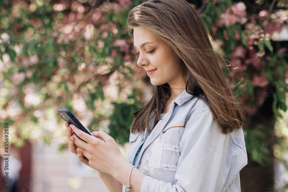 Happy hipster woman typing by mobile phone outdoors. Cheerful girl with smartphone in park on a background of sakura trees. Smiling lady holding cellphone in hands outside