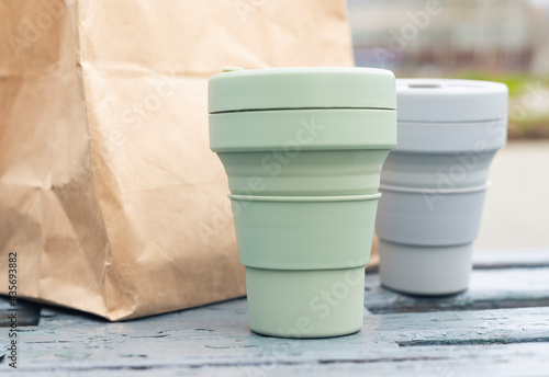 Two collapsible eco cups and craft parcel on city park bench. Reusable sustainable silicon mugs for takeaway tea or coffee.