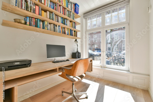 Modern interior of light study room with wooden wall mounted table and computer under shelves with books photo