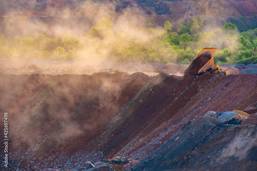 A dump truck unloads rock in a quarry. A dumper unloads stones in a opencast mine, forming huge clouds of dust, illuminated by sunlight.