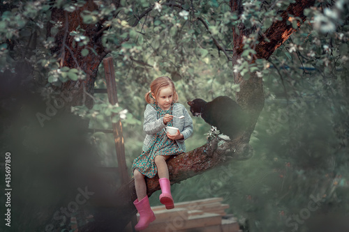 Funny little girl is trying to feed a black cat with a sour cream in a spoon sitting on the blossoming apple tree. Image with selective focus and toning