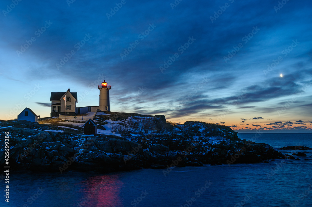 The moon begins to set over a cold snowy winter sunrise at the Nubble Lighthouse in York Maine