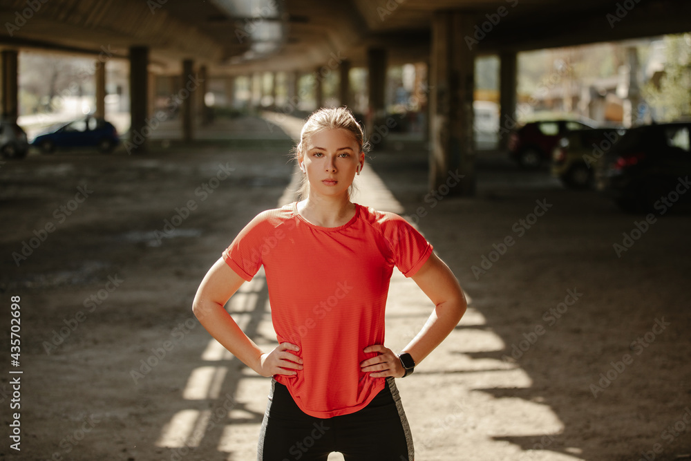 Blonde girl runner with earphones, looking at the camera, posing with hands on hips.