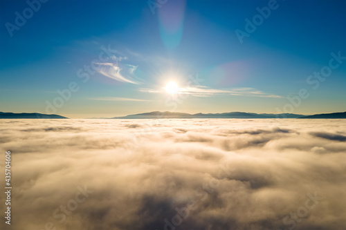 Aerial view of vibrant sunset over white dense clouds with distant dark mountains on horizon. © bilanol