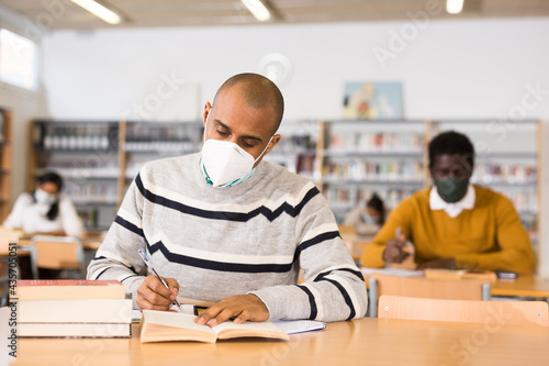 Focused Latin American adult wearing protective face mask looking for information in books in public library. New normal in coronavirus pandemic..
