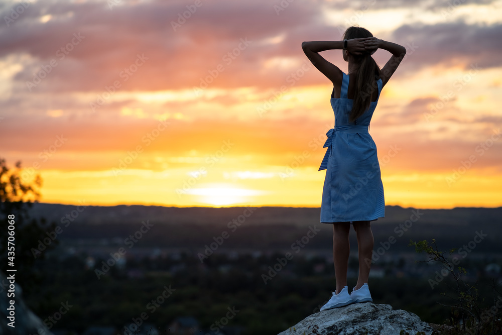 A young woman in summer dress standing outdoors enjoying view of bright yellow sunset.
