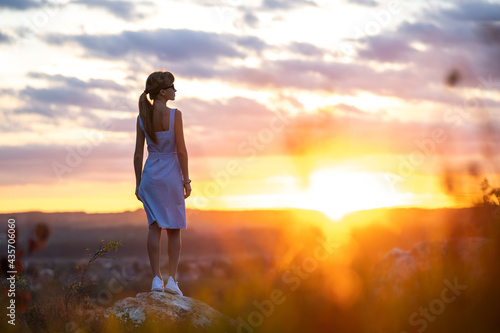 A young woman in summer dress standing outdoors enjoying view of bright yellow sunset.