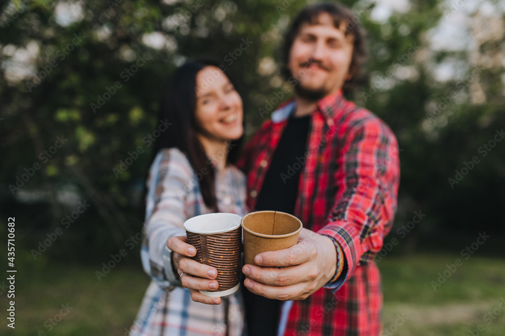 A handsome man and a girl in plaid shirts are resting in nature with hot coffee, tea in an eco-friendly paper cup in their hands. Photography, concept. A handsome man and a girl in plaid shirts are resting in nature with hot coffee, tea in an eco-friendly paper cup in their hands. Photography, concept.