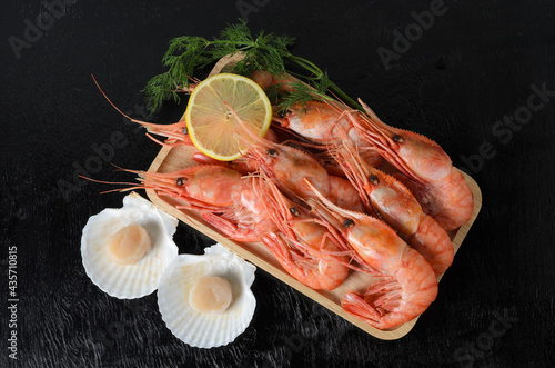 Fototapeta Naklejka Na Ścianę i Meble -  Boiled shrimps in a shell with dill and lemon in a wooden bowl with a raw scallop in a shell on a dark background.