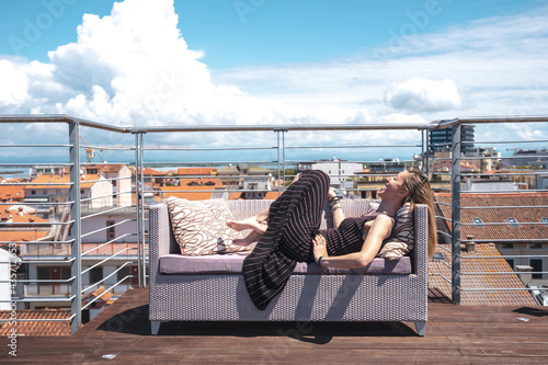 laughs girl lying in a sofa on the top floor terrace on a sunny day. on the background the roofs of the city