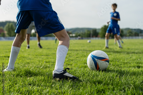 Fototapeta Naklejka Na Ścianę i Meble -  Close up on legs of unknown football soccer player caucasian boy child standing on the field in training or game by the ball kicking with unknown blurred players in background copy space