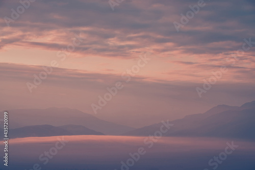 Scenic dawn mountain landscape with lilac low clouds in valley among mountains silhouettes under cloudy sky. Vivid sunset or sunrise scenery with low clouds in mountain valley in pink violet colors.