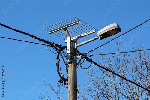Metal wire mesh stand prepared for stork nest mounted with strong metal pipes on top of dilapidated concrete utility pole with old street lamp and multiple connected thick black electrical wires surro
