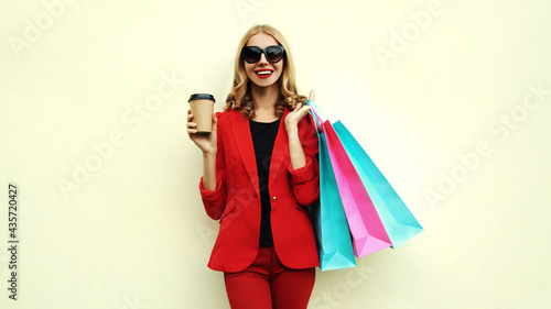 Portrait of happy smiling young woman with shopping bags and coffee cup wearing a red business blazer on a background