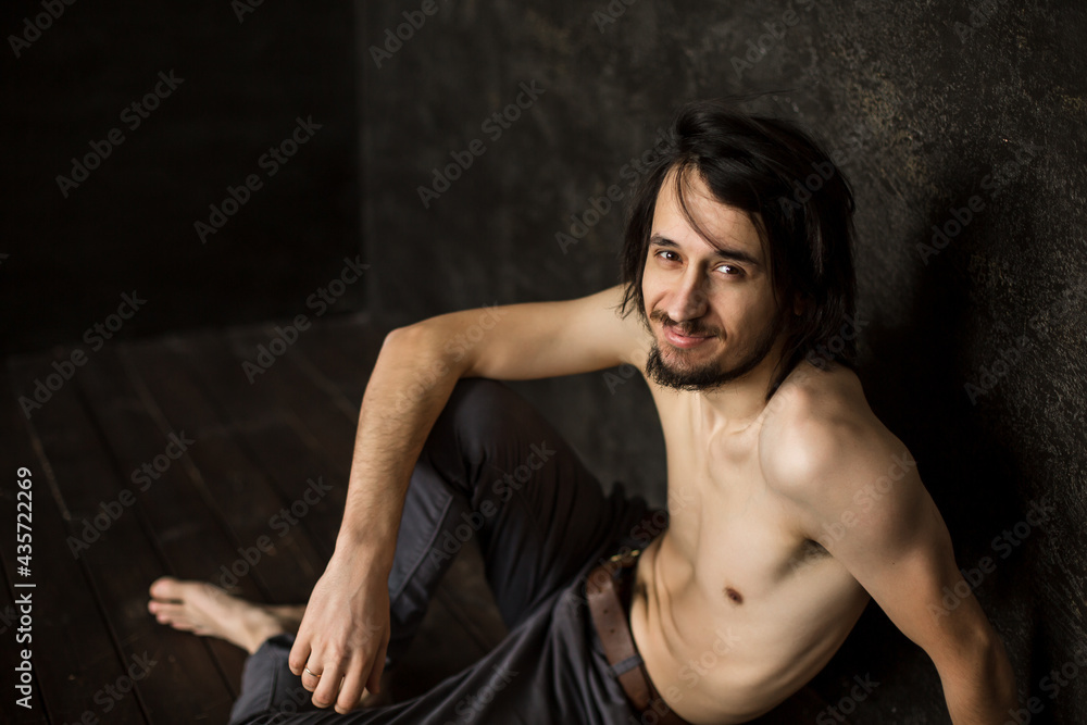 Skinny man without T-shirt sits looking at camera from below, beard mustache.