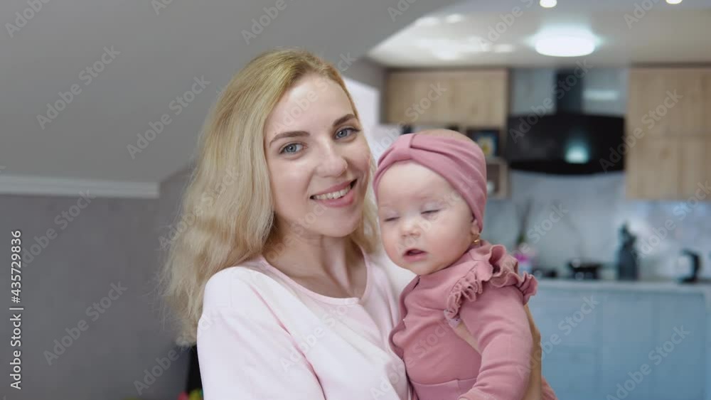 Mother holds baby in her arms on the background of a modern kitchen set and looks into the camera. Baby girl with blue eyes in a cute pink suit sits in mom's lap Mother holds baby in her arms on the background of a modern kitchen set and looks into the camera. Baby girl with blue eyes in a cute pink suit sits in mom's lap