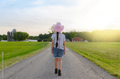 Girl walking on the road to the farm during the sunset surrounded by green grass and wearing a pink hat