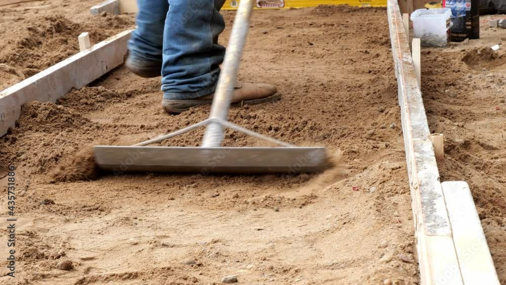 A rake tool is used by a working man wearing denim and boots to level dirt in preparation for a new cement concrete sidewalk, at a construction job site. A rake tool is used by a working man wearing denim and boots to level dirt in preparation for a new cement concrete sidewalk, at a construction job site.