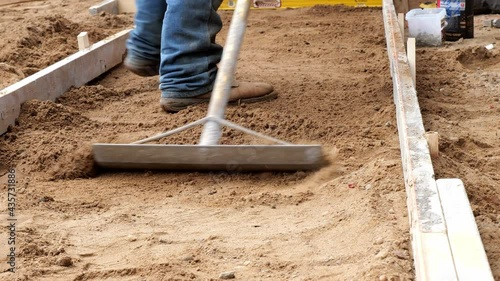 A rake tool is used by a working man wearing denim and boots to level dirt in preparation for a new cement concrete sidewalk, at a construction job site.