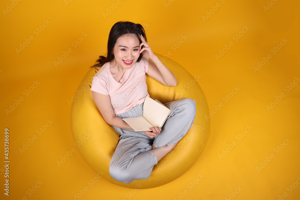 Beautiful young south east Asian woman sits on a yellow beanbag seat orange yellow color background pose fashion style elegant beauty mood expression rest relax think emotion read book top view Beautiful young south east Asian woman sits on a yellow beanbag seat orange yellow color background pose fashion style elegant beauty mood expression rest relax think emotion read book top view