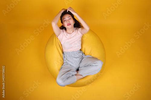 Beautiful young south east Asian woman sits on a yellow beanbag seat orange yellow color background pose fashion style elegant beauty mood expression rest relax think emotion top view © oqba